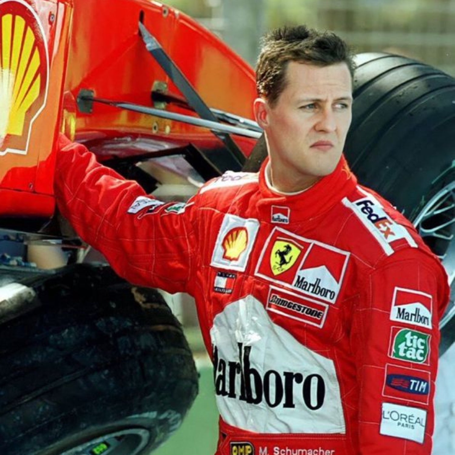 Schumacher in a red Ferrari F1 racing suit with various sponsor logos next to a Formula 1 car.