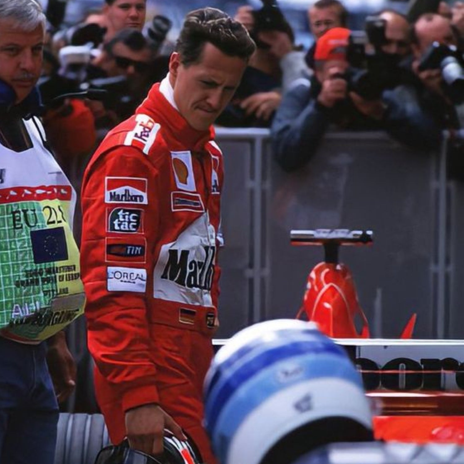 Michael Schumacher, in a Ferrari red F1 racing suit with various sponsor logos, standing in front of a Formula 1 car.
