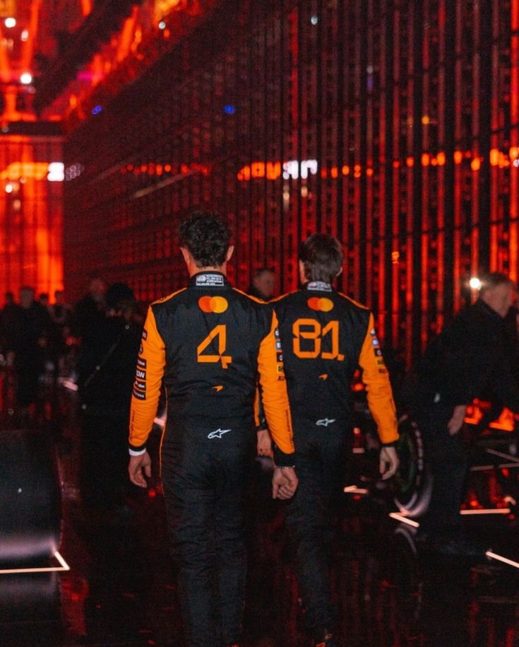 Oscar and Lando in McLaren 2025 F1 racing suits with numbers 4 and 81 standing in front of a vibrant, illuminated background.